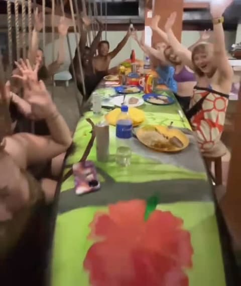 Guests cheering with their hands up around a long table painted with a giant red hibiscus, plates of food and drinks between them.