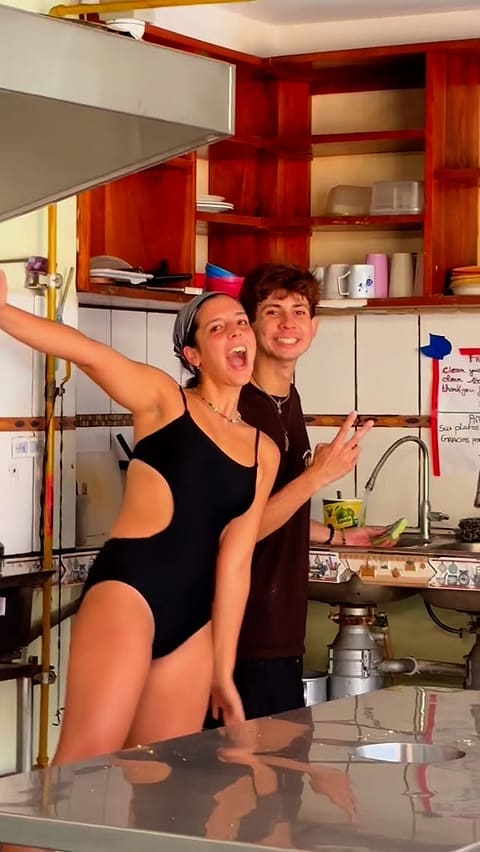 Two travellers laughing in the hostel's shared kitchen, one throwing a peace sign, with wooden shelves of mugs and plates behind them.