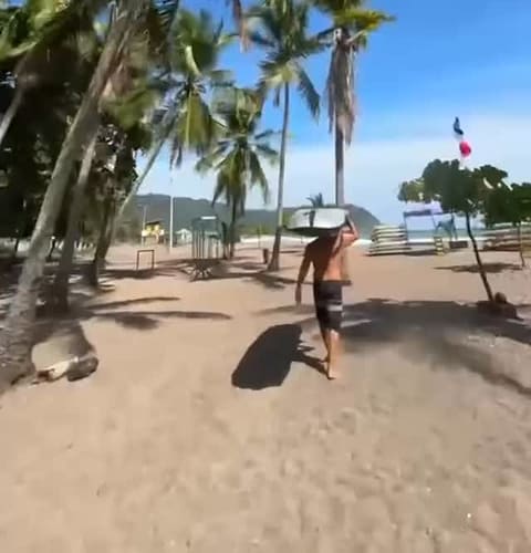 A surfer walks across the sand at Playa Jacó with his board balanced on his head, under tall palm trees.