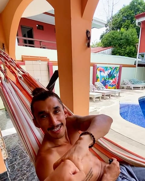 The pool courtyard at Hostel de Haan: orange arches, a striped hammock with a smiling guest, loungers, the toucan mural and the blue pool.