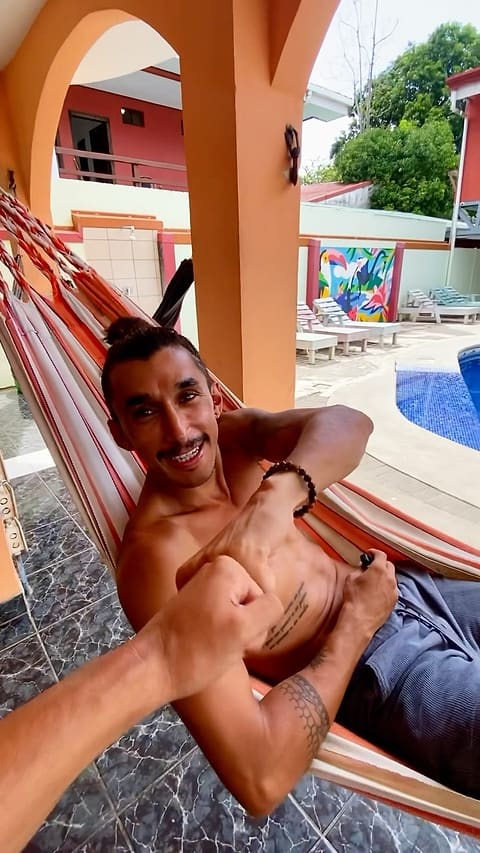 A guest grins from a striped orange hammock under the hostel's orange arches; behind him, white loungers, a colourful toucan mural and the blue-tiled pool.