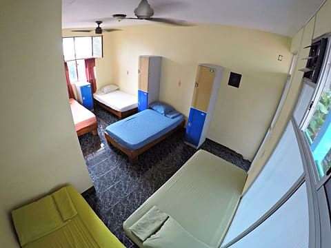 El dormitorio mixto de 5 camas: cinco camas individuales de colores, sin camarotes, lockers altos azules y dos abanicos de techo.