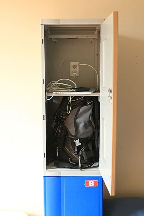 An open dorm locker with a backpack inside and a power outlet with charging cables on the shelf.