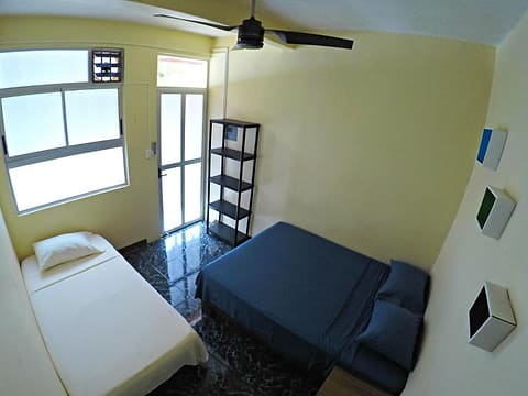 Habitación privada con una cama individual y una matrimonial con cobijas azul oscuro, abanico de techo, repisas y una ventana luminosa.