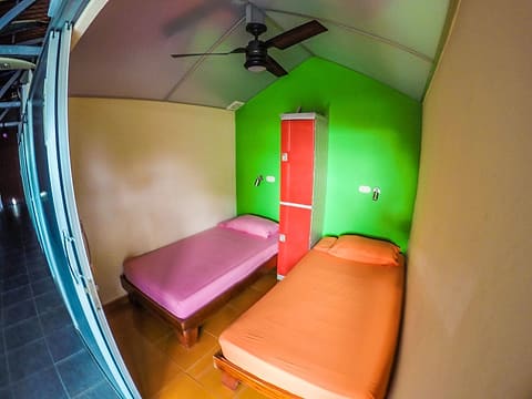 Room with two single beds in pink and orange, a red locker against a bright green wall and a ceiling fan.