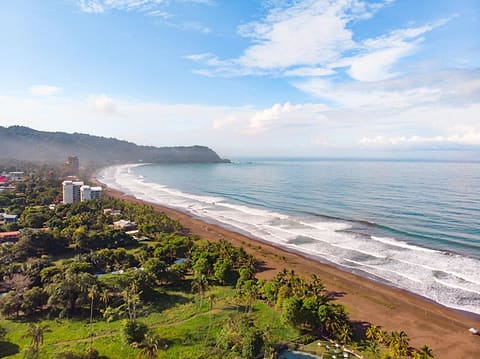 Panorama of the bay of Jacó: green hills, the long dark-sand beach and the town behind it.